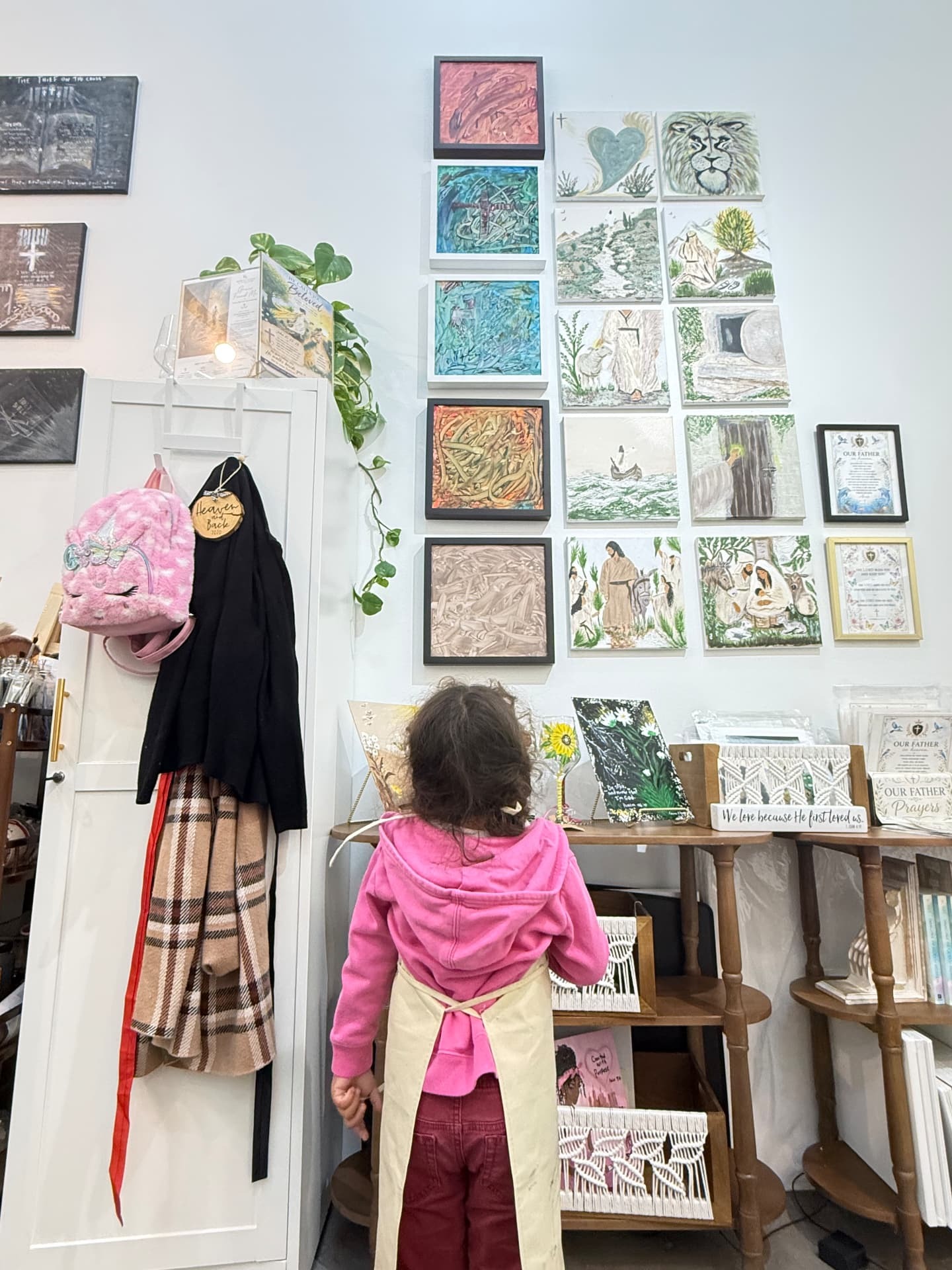 A four-year-old girl in a pink hoodie and painting apron, seen from behind, looking at a studio wall covered in colorful paintings